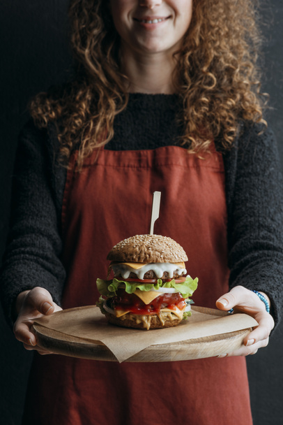 vista recortada de chica en delantal sosteniendo tablero de madera con hamburguesa de queso casera grande
 - Foto, Imagen