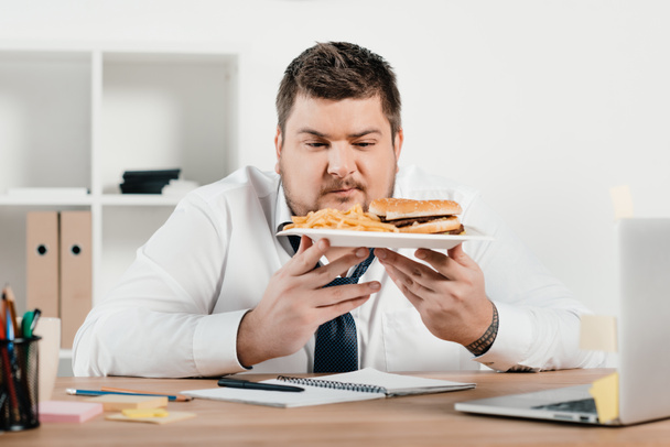 overweight businessman with hamburger and french fries in office - 写真・画像