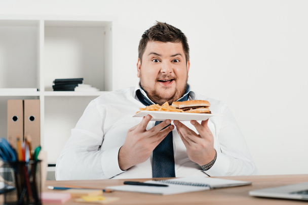 excited overweight businessman with hamburger and french fries in office - Photo, Image