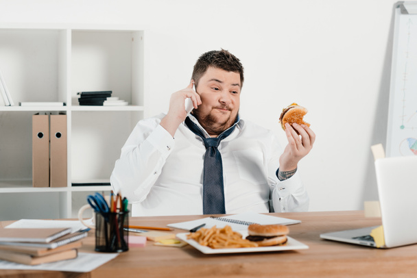 uomo d'affari in sovrappeso che parla sullo smartphone mentre mangia fast food in ufficio
 - Foto, immagini