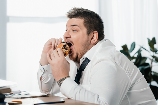 fat businessman talking on smartphone while eating hamburger in office - Photo, Image