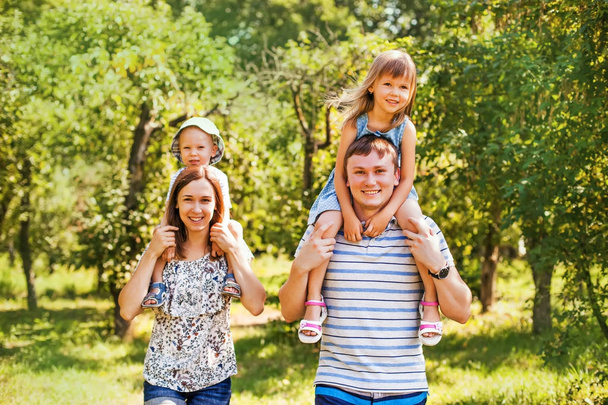 Uma família branca de quatro pessoas com crianças sentadas nos ombros dos pais desfrutando em um parque em um dia ensolarado
  - Foto, Imagem