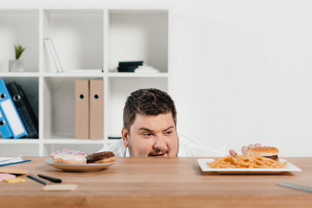 głodny biznesmen tłuszczu, wybierając pączki lub hamburger z frytkami na obiad - Zdjęcie, obraz
