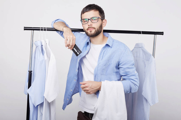 Portret van bebaarde jonge Europese man in stijlvolle bril kunststof creditcard op camera trots toont, gevoel opgewonden over winkelen, nieuwe aankopen, staan in de winkel onder de blauwe en witte kleding. - Foto, afbeelding