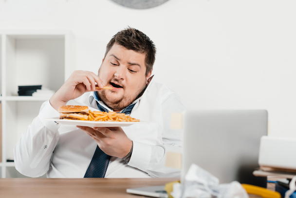overweight businessman having lunch with hamburger and french fries in office - 写真・画像