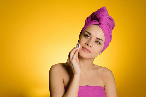 a young girl with a pink towel on her head looks after the skin on her face, cleans it with a cotton pad - Foto, Imagen