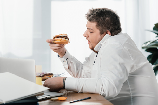 hombre de negocios con sobrepeso comiendo hamburguesa mientras trabaja en la oficina
 - Foto, imagen
