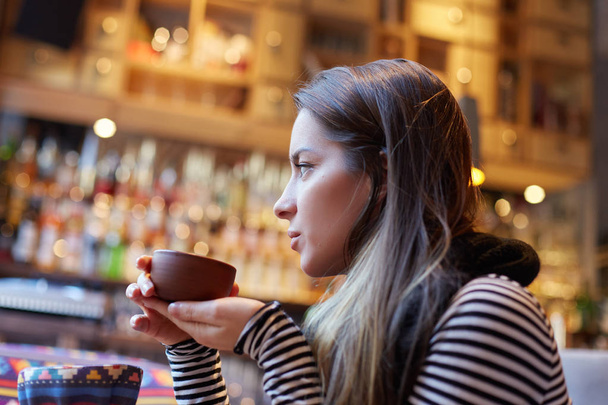Bild von gut aussehenden Teenager-Mädchen mit süßem Lächeln sitzt im Café, schaut aus dem Fenster, trinkt Heißgetränk, fühlt sich entspannt. Menschen, moderner Lebensstil, Freizeit und Entspannung. - Foto, Bild
