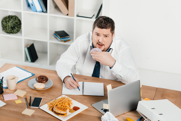 overweight businessman eating donuts, hamburger and french fries while wokring in office - Foto, Imagem