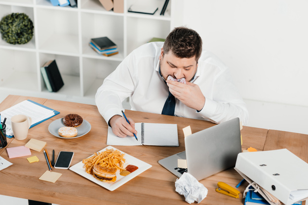 hombre de negocios con sobrepeso comiendo rosquillas, hamburguesas y papas fritas mientras se despierta en la oficina
 - Foto, imagen