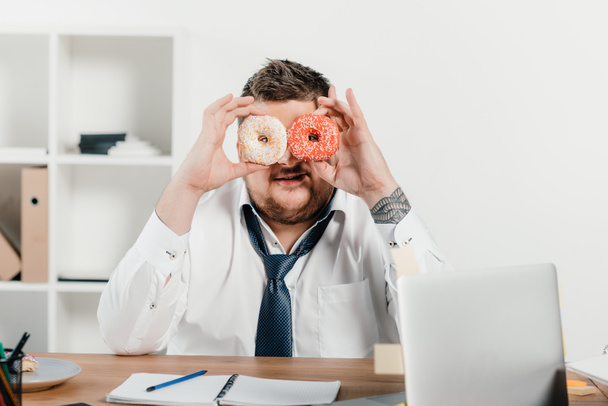 homem de negócios com sobrepeso segurando donuts na frente da cara no escritório
 - Foto, Imagem