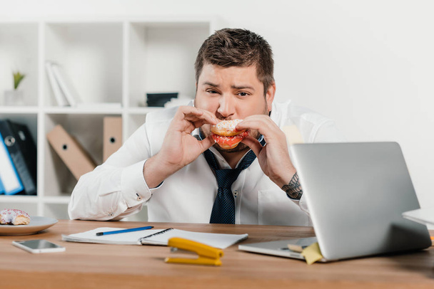 hombre de negocios con sobrepeso comiendo donas en el lugar de trabajo con computadora portátil
 - Foto, Imagen