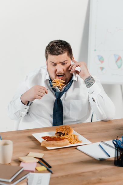 overweight businessman talking on smartphone while eating junk food at workplace - Photo, Image