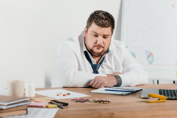 overweight businessman sitting at workplace and looking at pills - Φωτογραφία, εικόνα