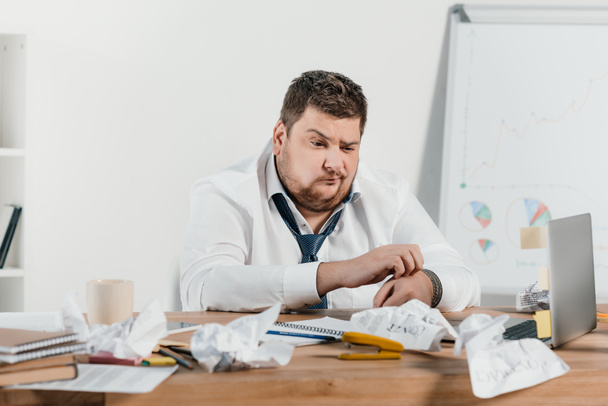 confundido hombre de negocios con sobrepeso sentado en el lugar de trabajo con papeles arrugados
 - Foto, Imagen
