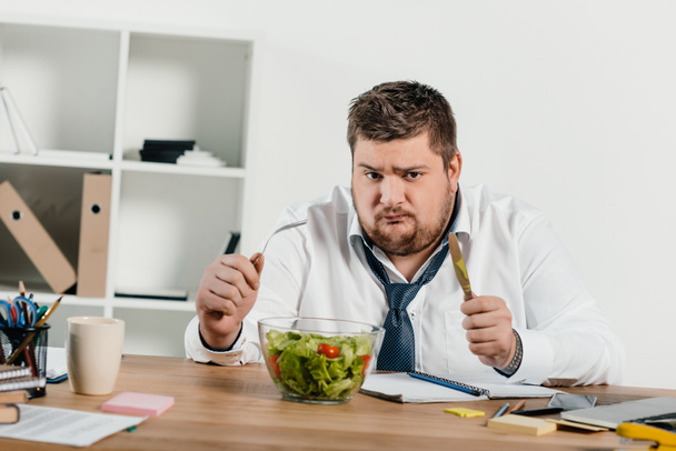 smutny nadwagą biznesmen z widelcem i nożem jedzenie świeże sałatki w miejscu pracy - Zdjęcie, obraz