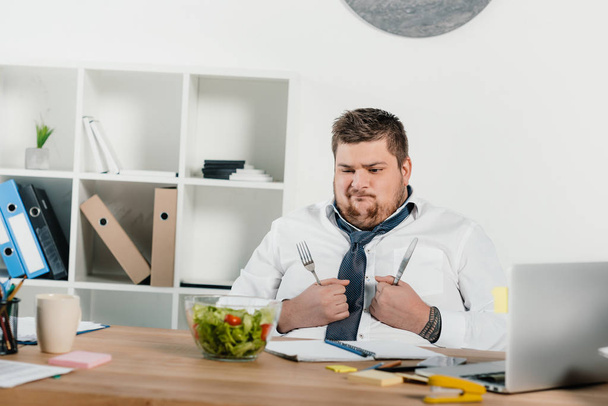 confundido hombre de negocios con sobrepeso con tenedor y cuchillo mirando ensalada fresca
 - Foto, imagen