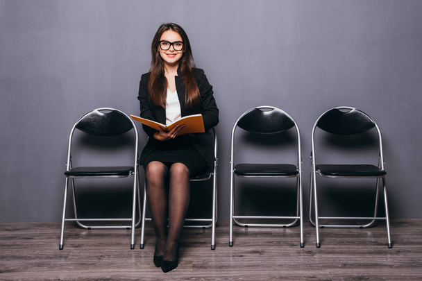 Устал ждать. Скучная молодая деловая женщина держит бумагу и смотрит в сторону, сидя на стуле на черном фоне
 - Фото, изображение