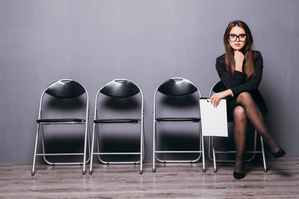 Aspetto un colloquio. Fiduciosa giovane donna d'affari in possesso di carta mentre seduto sulla sedia contro sfondo grigio
 - Foto, immagini