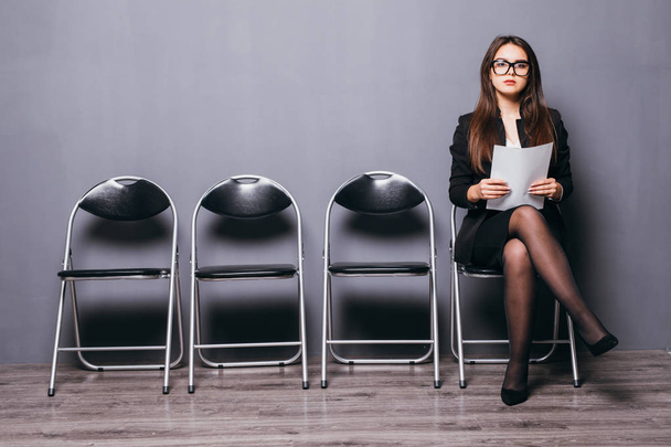 Чекаю на співбесіду. Впевнена молода бізнес-леді тримає папір, сидячи на стільці на сірому фоні
 - Фото, зображення