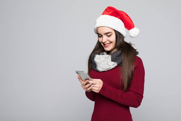 Portret brunetka, młoda kobieta w czerwonym Świętego Mikołaja za pomocą telefonu komórkowego, na białym tle na szarym tle - Zdjęcie, obraz