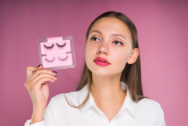 cute girl with big lips holding false eyelashes and looking away thoughtfully, isolated on pink background - 写真・画像