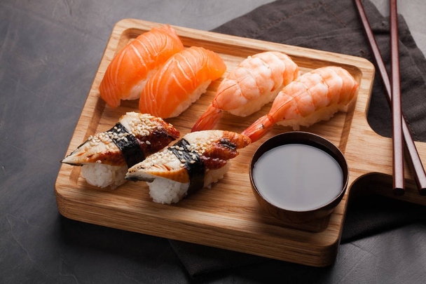 Zestaw sushi z łososiem, krewetkami i węgorz z miski sos sojowy i drewniane pałeczki do jedzenia na płycie. Pysznych potraw japońskich na ciemnym tle kamienia. Widok z góry - Zdjęcie, obraz