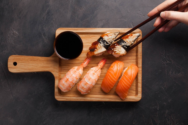 Un juego de sushi con salmón, camarones y anguila y palillos de madera en una tabla de madera. Deliciosa comida japonesa sobre un fondo de piedra oscura. La mano femenina toma los palillos en el sushi. Vista superior
 - Foto, imagen