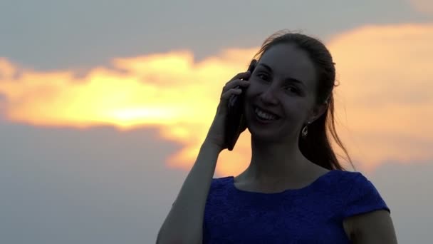 Μια νεαρή γυναίκα που χαμογελάει και να καλέσετε κάποιον από το smartphone στο ηλιοβασίλεμα - Πλάνα, βίντεο