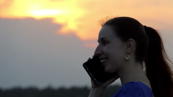 Una joven sonríe y llama a su amiga con su smartphone al atardecer
 - Metraje, vídeo