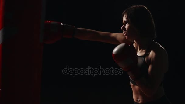 Sportieve brunette boksen sportschool biedt individuele training op stroke techniek. Voert strikes op een bokszak op een zwarte achtergrond. - Video