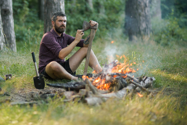 Man lumberjack with beard sharpen axe at bonfire - Foto, afbeelding