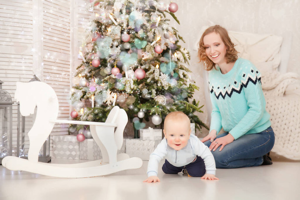 Pretty Slim Mother Wearing Bright Knitted Sweater and Blue Jeans Playing With Her Little Baby Near The Decorated Christmas Tree with New Year Gift Boxes. Real Family. - Фото, зображення