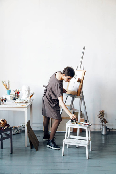 Female artist in her spacious white studio working with watercolor painting dipping the brush into glass with water.  Natural lighting. Disclosure of creativity concept. Vertical composition with copy space - Fotografie, Obrázek