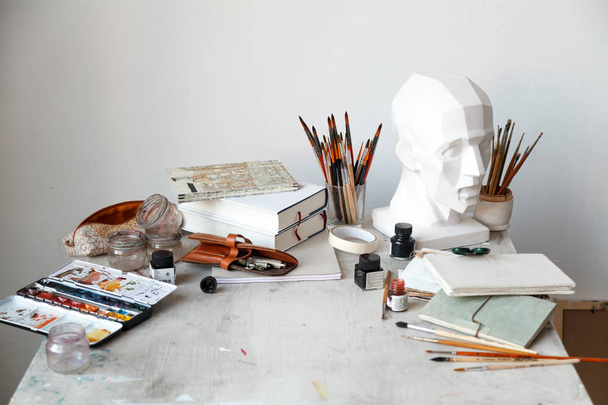 Interior of painting studio of freelance artist. Gypsum head, brushes, pencills, ink and paint bottles with sketchbooks on the table. Freelance artist lifestyle concept. Copy space - Foto, imagen