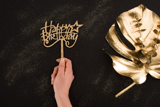 tiro cortado de mão feminina segurando sinal de aniversário feliz com folha de ouro deitado perto por
 - Foto, Imagem