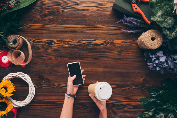 cropped image of florist holding disposable coffee cup and smartphone in hands  - Foto, imagen