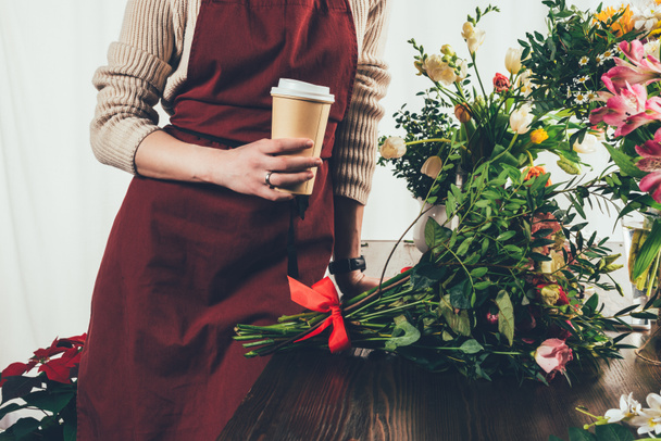 cropped image of florist holding coffee in paper cup and leaning on working table - Fotó, kép