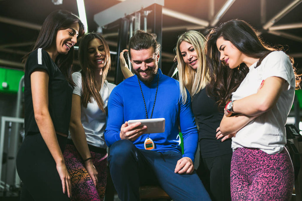 bonito personal trainer usando tablet digital, grupo de mulheres desportivas em torno dele no ginásio
  - Foto, Imagem