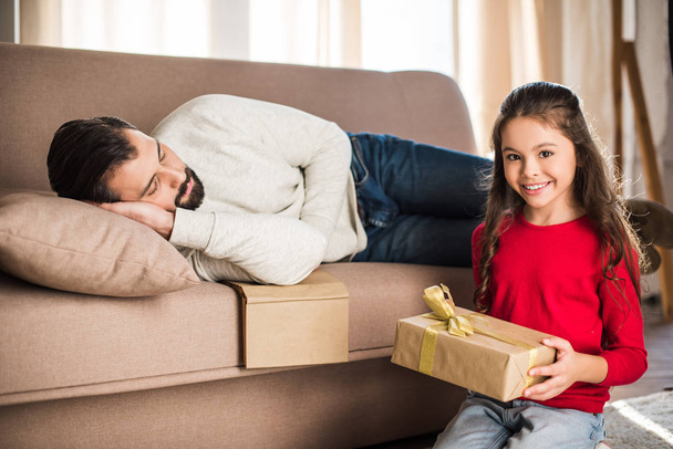 daughter holding gift box while father sleeping on sofa  - Valokuva, kuva
