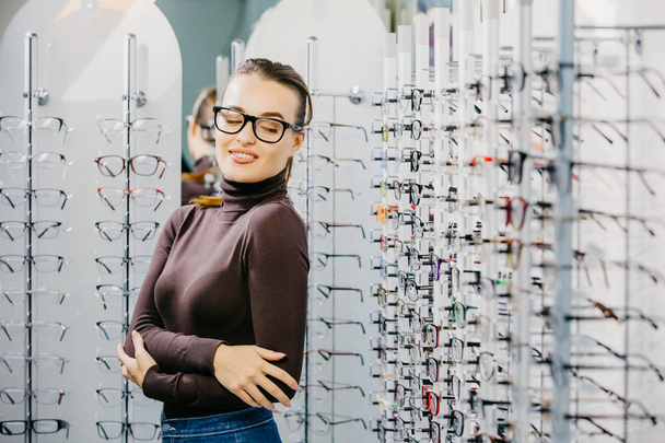 Ophthalmology. Young woman is choosing a glasses in optician store. Eyesight correction. - Фото, изображение
