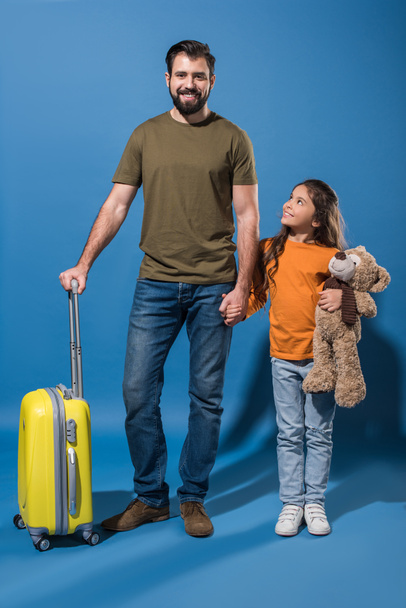 father and daughter standing with yellow travel bag and teddy bear on blue - Foto, Imagen
