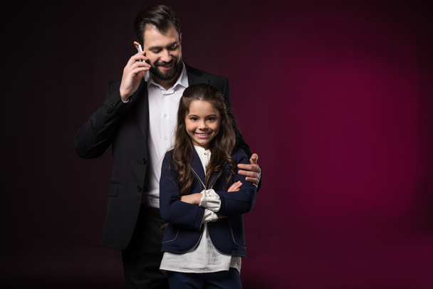 father talking by smartphone and hugging smiling daughter with crossed hands on burgundy - Valokuva, kuva