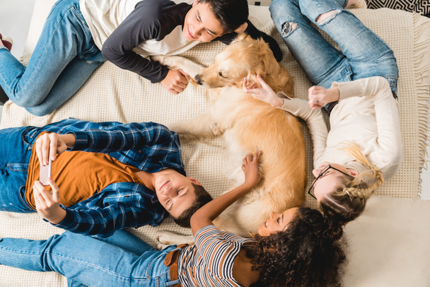overhead view of multiethnic teenagers lying on bed and taking selfie with dog - Photo, Image