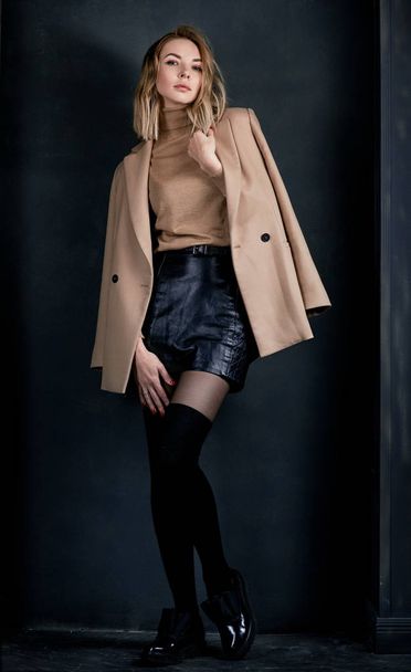 Retrato de joven atractiva mujer rubia bonita en la chaqueta beige y camisa de cuero, polainas, zapatos de charol en el fondo oscuro
 - Foto, Imagen