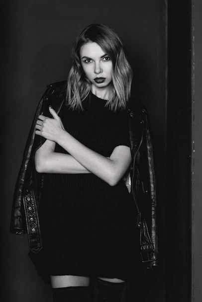 Retrato en blanco y negro de la joven atractiva mujer rubia con estilo en vestido corto, botas hasta la rodilla y chaqueta negra en la habitación minimalista con paredes oscuras en el estudio de fotografía
 - Foto, imagen