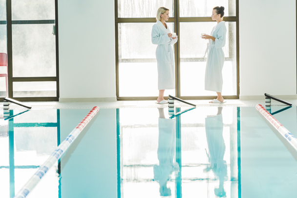 beautiful young women standing next to swimming pool at spa center and talking with coffee cups in hands - Photo, Image