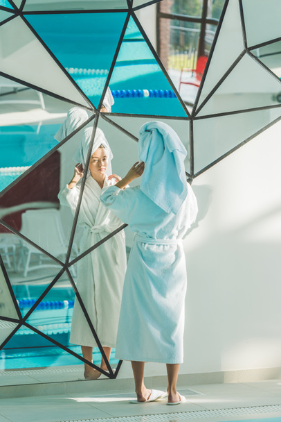 νεαρή γυναίκα στο μπουρνούζι και πετσέτα κοιτάζοντας καθρέφτη στο κέντρο ευεξίας - Φωτογραφία, εικόνα