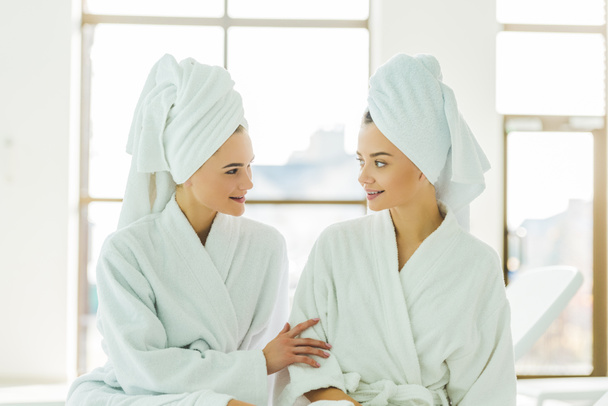 beautiful young women in bathrobes and towels on head talking to each other at spa center - Фото, изображение