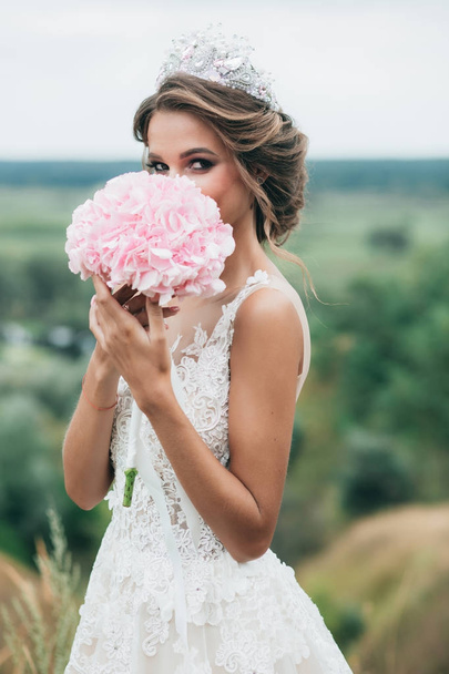The bride is holding a wedding bouquet of hydrangeas and stands on a hill overlooking a beautiful nature. Closes his face with a wedding bouquet. - Fotó, kép